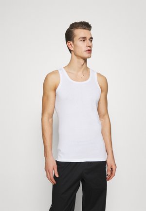 Hvid bomulds tanktop med rund halsudskæring og brede skulderstropper, båret med sorte bukser, der har en glat overflade.