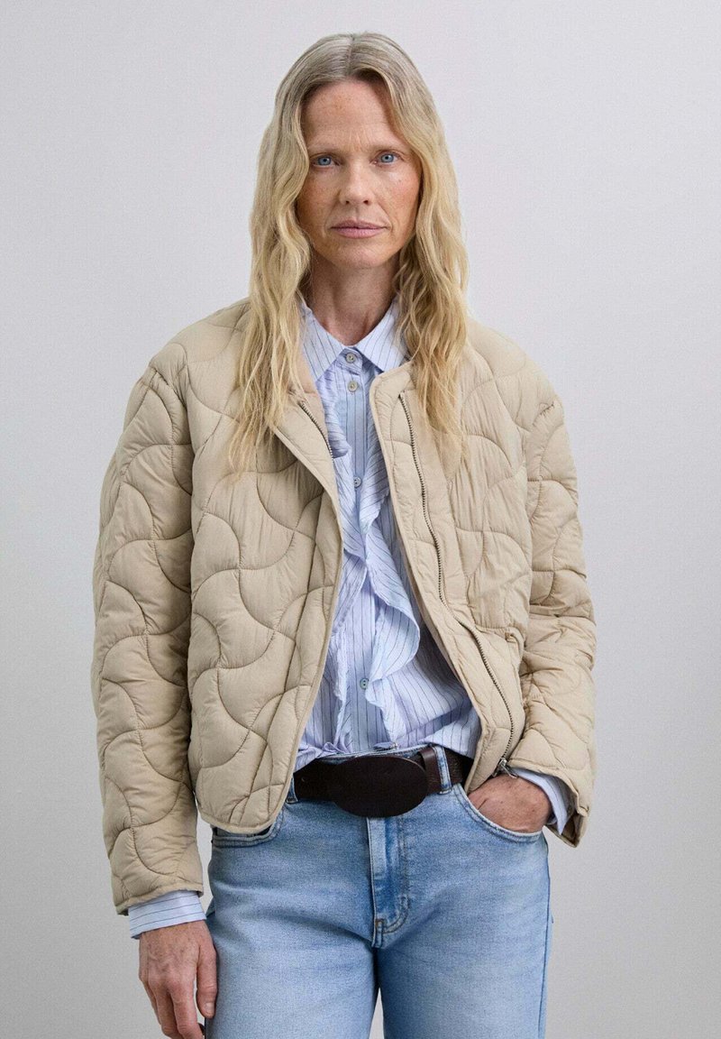 Chaqueta acolchada color beige con un corte relajado, que presenta un cuello redondo y costuras visibles. Combinada con una camisa a rayas de color azul claro y denim.