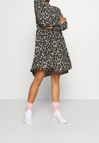 Blumenkleid mit langen Ärmeln, dunklem grünen Grundton, rosa und weißen Blumen, hohem und niedrigem Saum, kombiniert mit weißen Sneakers und rosa gestreiften Socken.