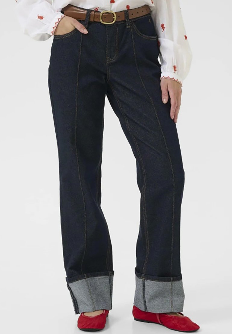 Person trägt dunkelblaue Jeans mit umgeschlagenen Säumen, braunen Gürtel, weiße Bluse mit roter Stickerei und rote flache Schuhe, steht mit einer Hand in der Tasche.