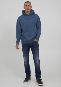 Blå bomullshoodie med känguruficka och tryck på framsidan, kombinerad med mörk denimjeans med en urblekt finish, bärs med svarta sneakers.