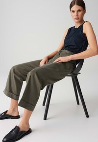 Black sleeveless top, olive green wide-leg trousers, rolled cuffs, black slip-on shoes, sitting on a black chair against a neutral background.