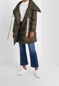 Olivgrön vadderad jacka med stor huva, vita dragsnören och dragkedja. Bärs över en vit topp och blå utsvängda jeans, kombinerad med vita sneakers.