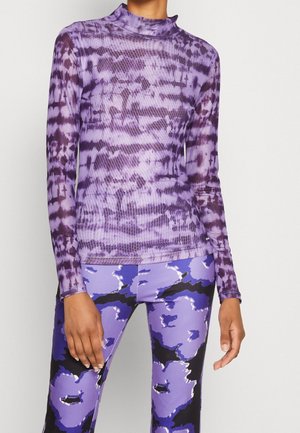 Kobieta ubrana w długi rękaw fioletowej koszulki w stylu tie-dye i legginsy z wysokim stanem o abstrakcyjnym wzorze w odcieniach fioletu, czerni i bieli, stojąca na tle jednolitego tła.