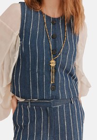 Gilet di denim con strisce bianche verticali, quattro bottoni neri e scollatura rotonda. Sovrapposto a una blusa trasparente a maniche puff e collana dorata.