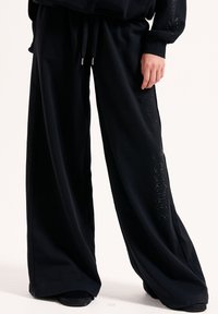 Schwarze weite Sweatpants mit einem Kordelzugbund, ausgestattet mit einem glänzenden Logodetail entlang der linken Seite, aus weichem, bequemem Stoff gefertigt.
