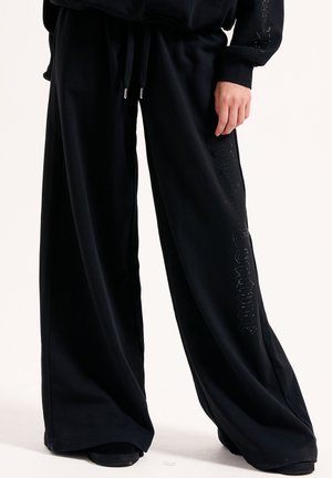 Schwarze weite Sweatpants mit einem Kordelzugbund, ausgestattet mit einem glänzenden Logodetail entlang der linken Seite, aus weichem, bequemem Stoff gefertigt.