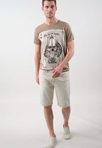 T-shirt graphique beige avec un design de crâne et des éléments de cartes à jouer. Associé à un short en denim de couleur claire et des chaussures slip-on légères.