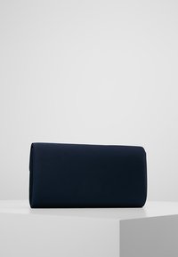 Obdélníková kabelka v námořnické modři s hladkým povrchem, postavená vzpřímeně na bílém podstavci na jednobarevném světle šedém pozadí.