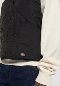 Chaleco acolchado negro con botones de presión, bolsillo frontal y un parche de logo. Llevado sobre un suéter crema, con puños acanalados y jeans de mezclilla.