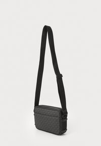 Sac bandoulière noir en tissu à motifs arborant la lettre "M". Forme rectangulaire, bandoulière ajustable et fermeture éclair.