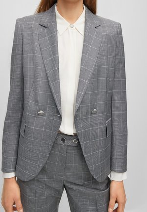 Frau trägt grauen karierten doppelt geknöpften Blazer und passende Hose über einer weißen, geknöpften Bluse vor neutralem Hintergrund.