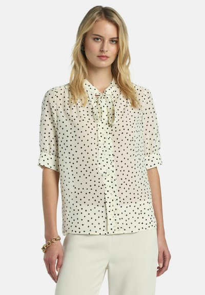 Krém színű blúz fekete pöttyökkel, masnis nyakkal, rövid felhajtott ujjakkal és gombos elülső résszel. Áttetsző anyag enyhén texturált felülettel.