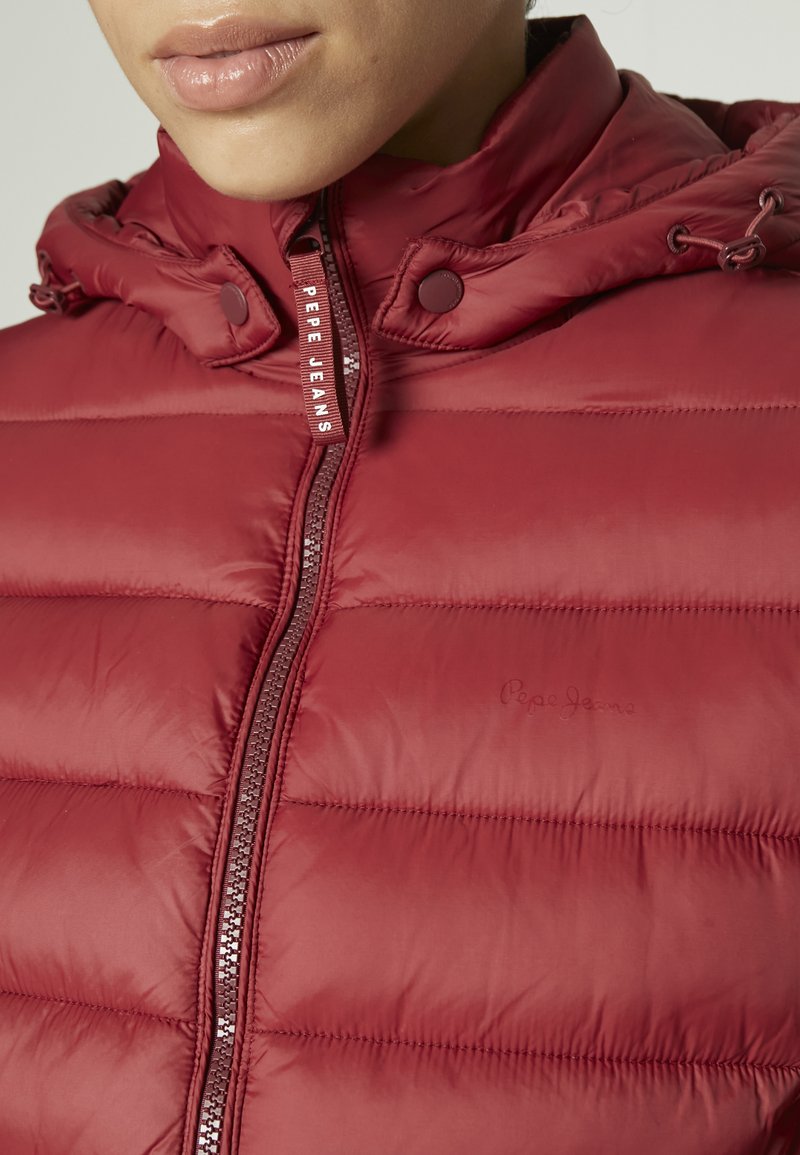 Chaqueta acolchada roja con capucha, que presenta secciones acolchadas, un cierre frontal y una etiqueta de logo. Textura lisa y brillante con cordones en la capucha.