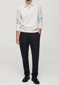 Vaaleanharmaa paita, jossa on kaulus ja kolme nappia, yhdistettynä mustiin suippeneviin housuihin, joissa on kaksi etutaskua. Mustat nauhakengät.