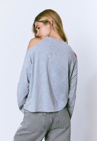 Sudadera gris, ligera, con un diseño de hombro descubierto. Presenta un corte relajado y una textura suave, combinada con pantalones de mezclilla grises.