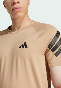 Light brown athletic t-shirt with a round neckline, short sleeves, and black accent stripes featuring perforated patterns and a logo on the chest.