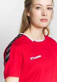 Jeune femme à la peau claire et aux cheveux blonds porte un maillot de sport rouge Hummel avec des bandes chevrons noires et grises sur l'épaule.