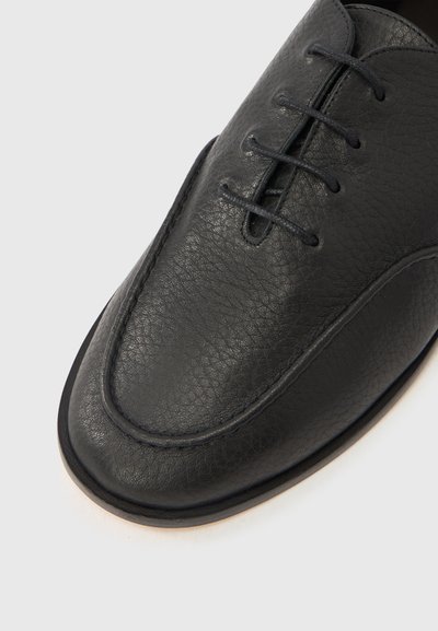 Μαύρο δερμάτινο παπούτσι με υφασμάτινη επιφάνεια, στρογγυλεμένη μύτη και τέσσερις οπές για κορδόνια. Διακριτικές λεπτομέρειες ραφής γύρω από την άκρη.