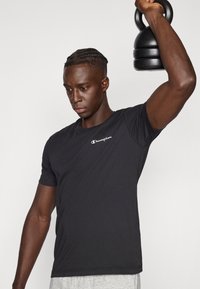 Μαύρο κοντομάνικο μπλουζάκι με διακριτικό λευκό λογότυπο, συνδυασμένο με ανοιχτό γκρι σορτς· το άτομο σηκώνει μια μαύρη αλτήρα kettlebell.