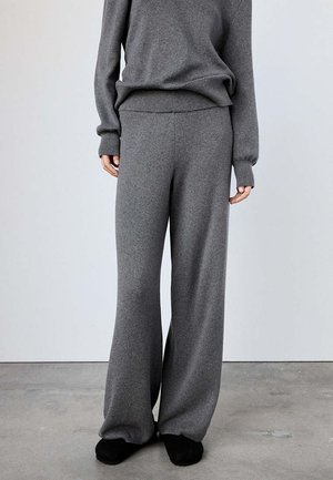 Personne portant un pantalon en maille gris ample et un pull gris assorti, debout sur un sol en béton contre un mur uni.