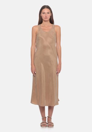 Donna vista frontale che indossa un vestito midi senza maniche in raso beige e sandali beige con cinturini di perle su uno sfondo chiaro e uniforme.