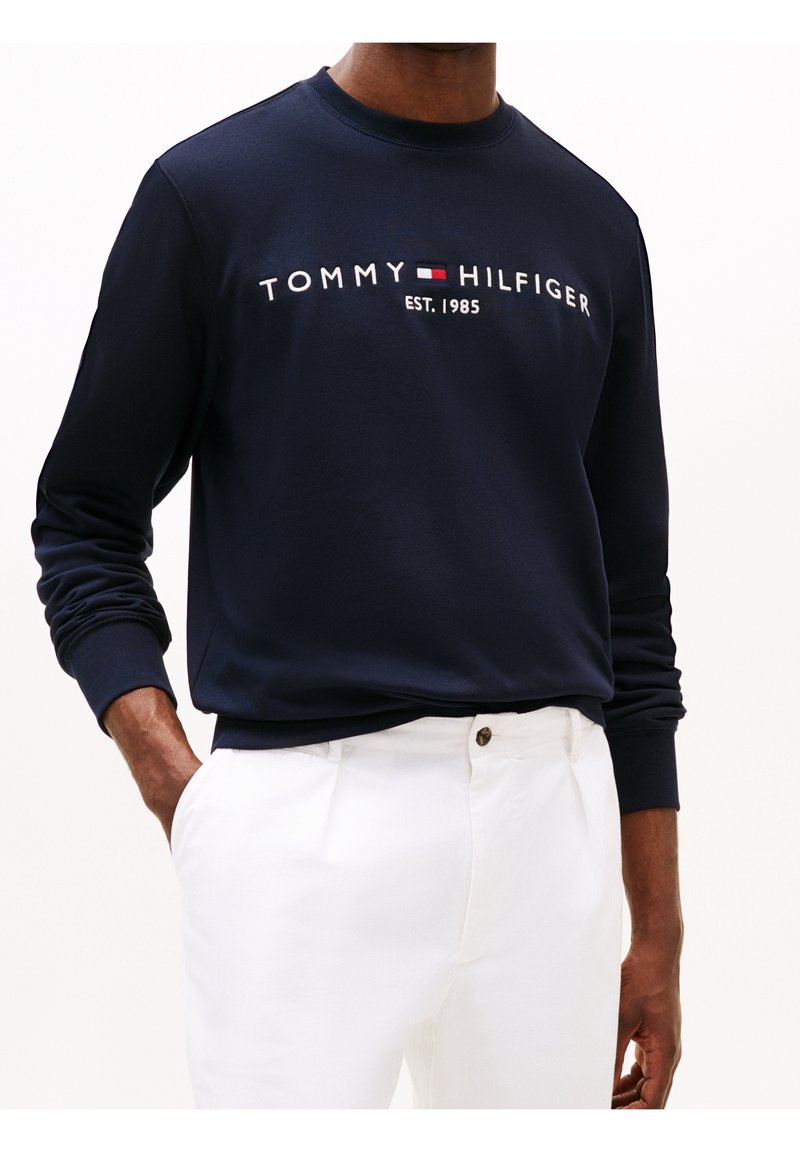 Homme portant un sweat-shirt Tommy Hilfiger bleu marine avec logo et "EST. 1985," associé à un pantalon blanc, mains partiellement dans les poches.