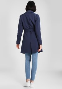 Femme avec des cheveux courts et foncés, portant un manteau bleu marine ceinturé, un jean skinny bleu clair et des baskets blanches, se tenant de dos sur un fond neutre.