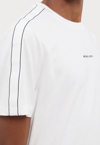 Tricou alb din bumbac cu guler rotund, având cusături decorative negre pe umeri și un mic logo pe piept.