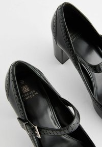 Zapatos de tacón alto de cuero negro con tira estilo Mary Jane, que presentan acentos perforados y un acabado texturizado. Incluye un detalle de hebilla de metal.