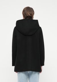 Person wearing a black hooded coat viewed from behind, paired with light blue jeans against a plain white background.