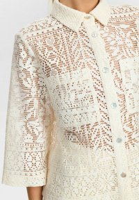 Spitzenhemd in Creme mit einem Hemdkragen, das über filigrane Muster, zwei aufgesetzte Taschen und perlenartige Knöpfe verfügt. Texturiertes Gewebe.