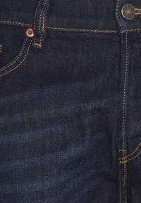 Granatowe dżinsy cechują się gładką fakturą, mosiężnymi akcentami i pomarańczowymi szwami wzdłuż szwów i kieszeni.