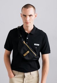 Juodas polo marškinėliai su baltu "Moschino" logotipu, derinami su juoda liemene, turinčia auksinį užtrauktuką ir užrašą "DIRECTION". Neutralios smėlio spalvos kelnės.