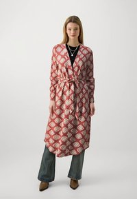 Lang, mønstret kappe i rustfarge med hvite blomsterdetaljer, med knytt belte i livet, lange ermer og en avslappet passform over mørke jeans.