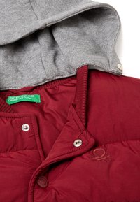 Červená zateplená bunda s šedou kapucí, ozdobená kovovými patentkami a zelenou nálepkou. Hladký povrch s jemným detailním logem na látce.