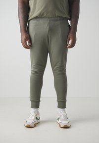 Olivgrüne Jogginghosen mit elastischem Bund und umgefalteten Knöcheln, kombiniert mit weißen Sneakers mit grünen Akzenten und strukturierten Sohlen.