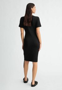 Black, fitted dress with short sleeves, knee-length, smooth texture, and side gathering details. Paired with black slip-on footwear.