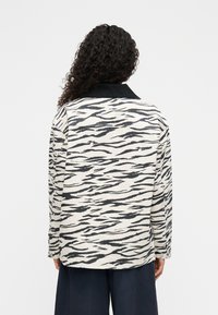 Πουκάμισο με μαύρες και λευκές ρίγες από υφασμάτινη υφή. Διαθέτει μαύρο γιακά και μακριά μανίκια, με στρογγυλεμένο τελείωμα και διακριτική επωνυμία.