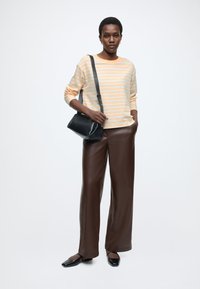 Pulover dungi galbene și gri, pantaloni maro din piele cu croială largă, geantă neagră purtată în diagonală și pantofi negri comozi. Texturi netede și croială lejeră.