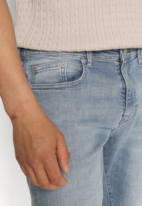 Jeans in denim azzurro chiaro con una vestibilità a vita media, cinque tasche e hardware con bottoni argentati. Il tessuto presenta leggeri sbiadimenti e una texture liscia.