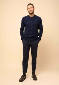 Cardigán azul marino con botones, combinado con pantalones oscuros de corte ajustado y zapatos de cuero marrones con cordones. El conjunto presenta un tejido texturizado y una silueta entallada.