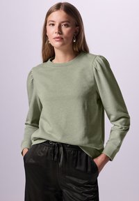 Ung kvinna som bär en ljusgrön långärmad topp med puffärmar och svarta byxor med dragsnöre, stående med händerna i fickorna mot en enfärgad bakgrund.