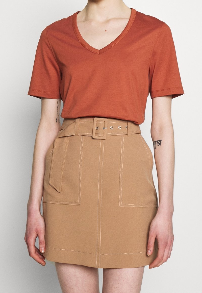 Personne portant un haut à manches courtes col en V couleur rouille, rentré dans une jupe beige taille haute avec une ceinture et des poches plaquées à l'avant.