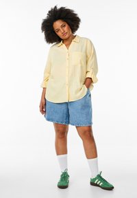 Ljust gult skjorta med knappar och bröstficka, tillsammans med denimshorts. Gröna sneakers och vita strumpor fulländar outfiten.
