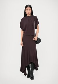 Tumma violetti mekko, jossa on virtaava siluetti, korkea kaula-aukko ja hulmuavat hihat, yhdistettynä polven ylle ulottuviin mustiin saappaisiin.