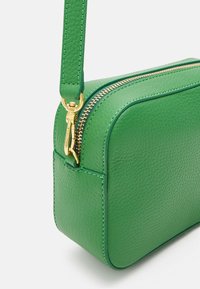 Borsa a tracolla in pelle verde con zip dorata visibile e gancio dorato che attacca la tracolla al corpo della borsa.