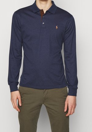 Homme portant un polo bleu marine à manches longues avec un col marron et un pantalon vert, debout devant un fond clair uni.