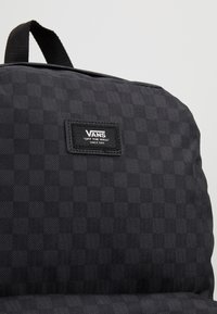Sac à dos noir avec un motif à damier texturé, une poignée supérieure et une étiquette rectangulaire avec le logo "VANS" cousue à l'avant.