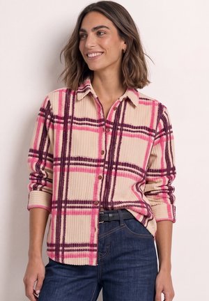 Jeune femme souriante portant une chemise à carreaux beige et rose boutonnée, manches retroussées, et un jean bleu foncé avec une ceinture noire.
